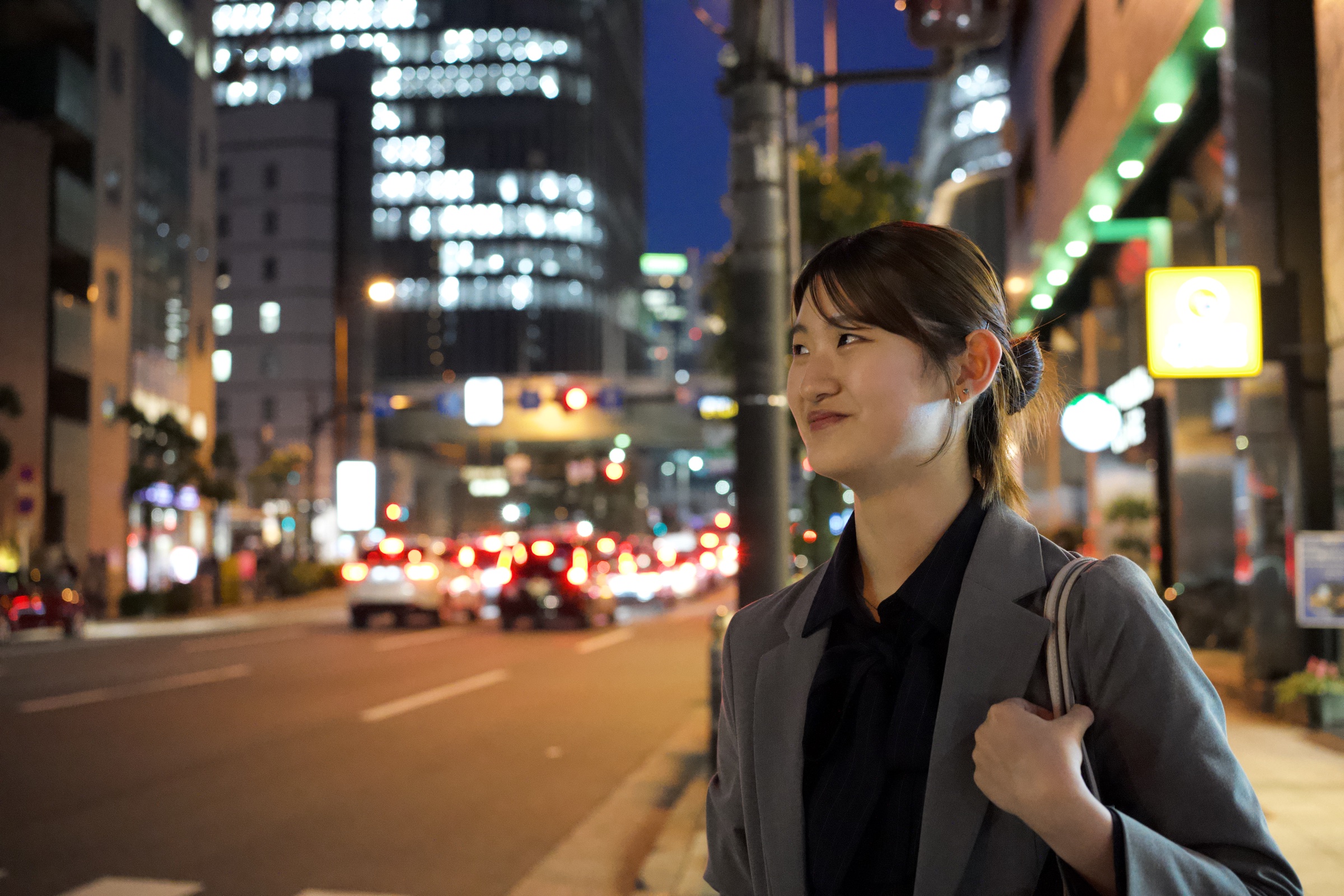 Young woman in a grey blazer and low bun looks away from the camera on an Osaka street at night, city lights and tail lights bokeh behind her, a subtle smile she's not giving you
