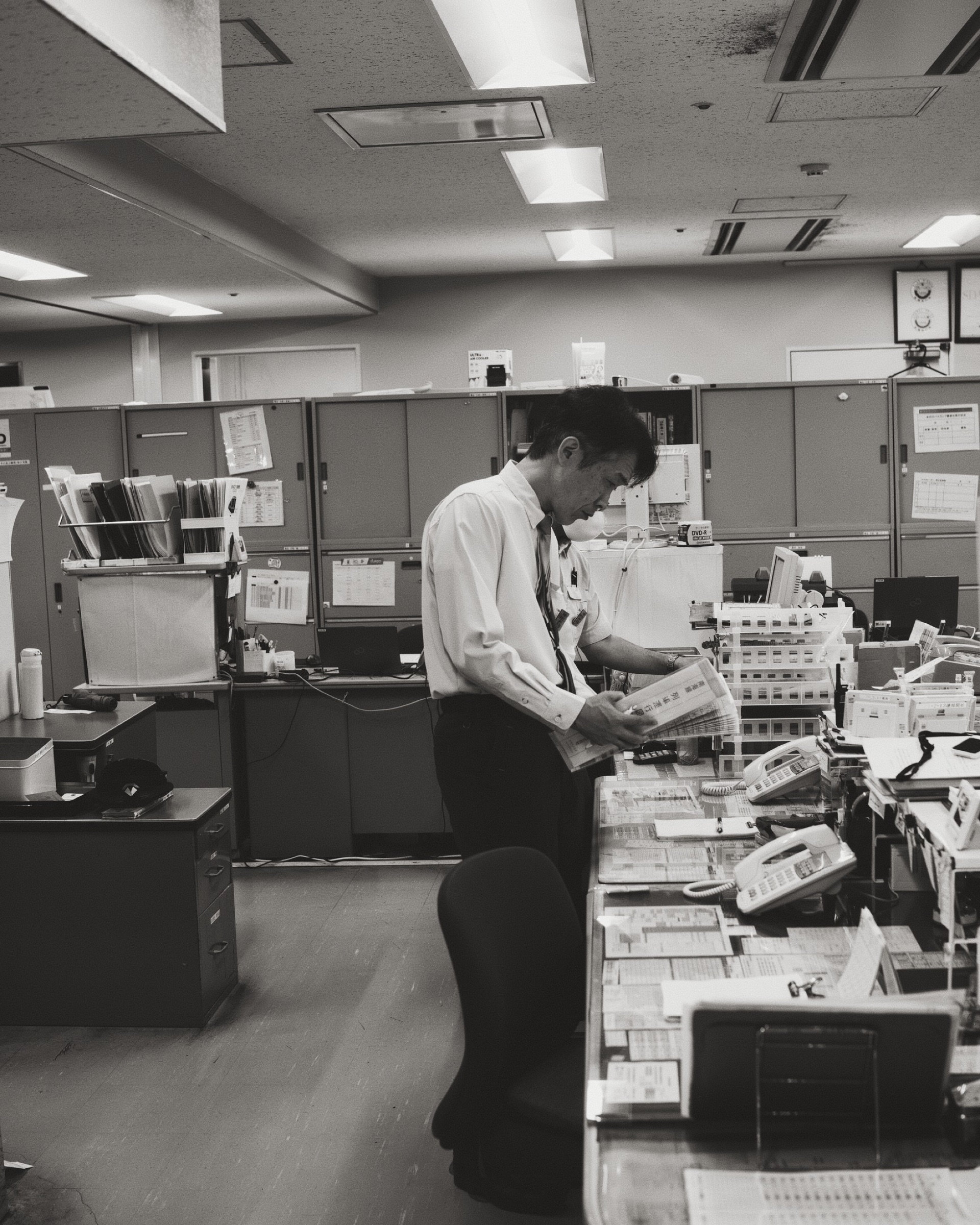 Japanese salaryman alone in a fluorescent-lit office reading documents after hours, black and white, quiet exhausted dignity