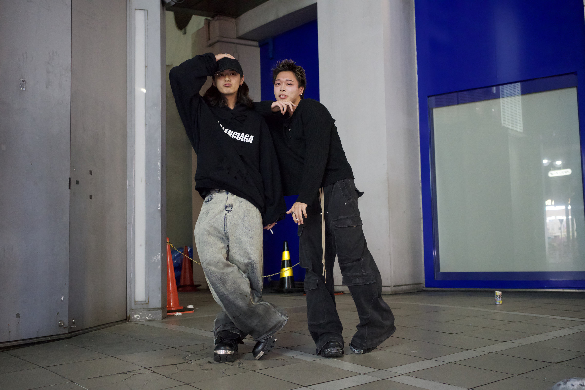Two young men pose against a blue-lit storefront in Tokyo at night, one in a Balenciaga hoodie leaning on the other