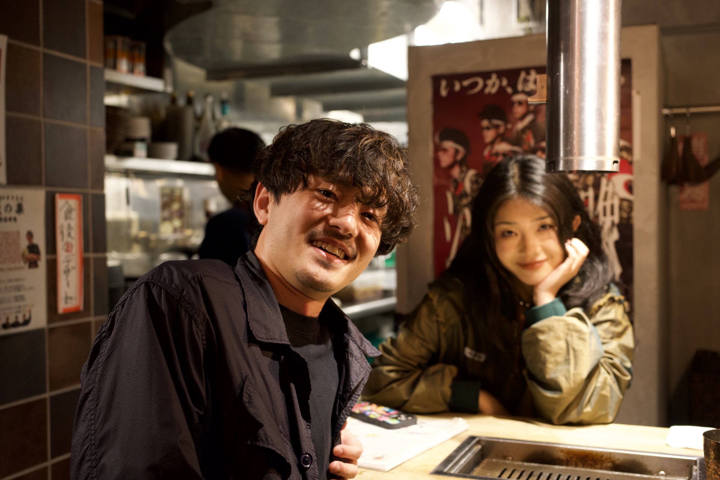 Man in black smiles warmly at the camera from behind a small Osaka restaurant counter, a woman with chin in hand rests behind him, warm kitchen light and a red anime poster on the tiled wall