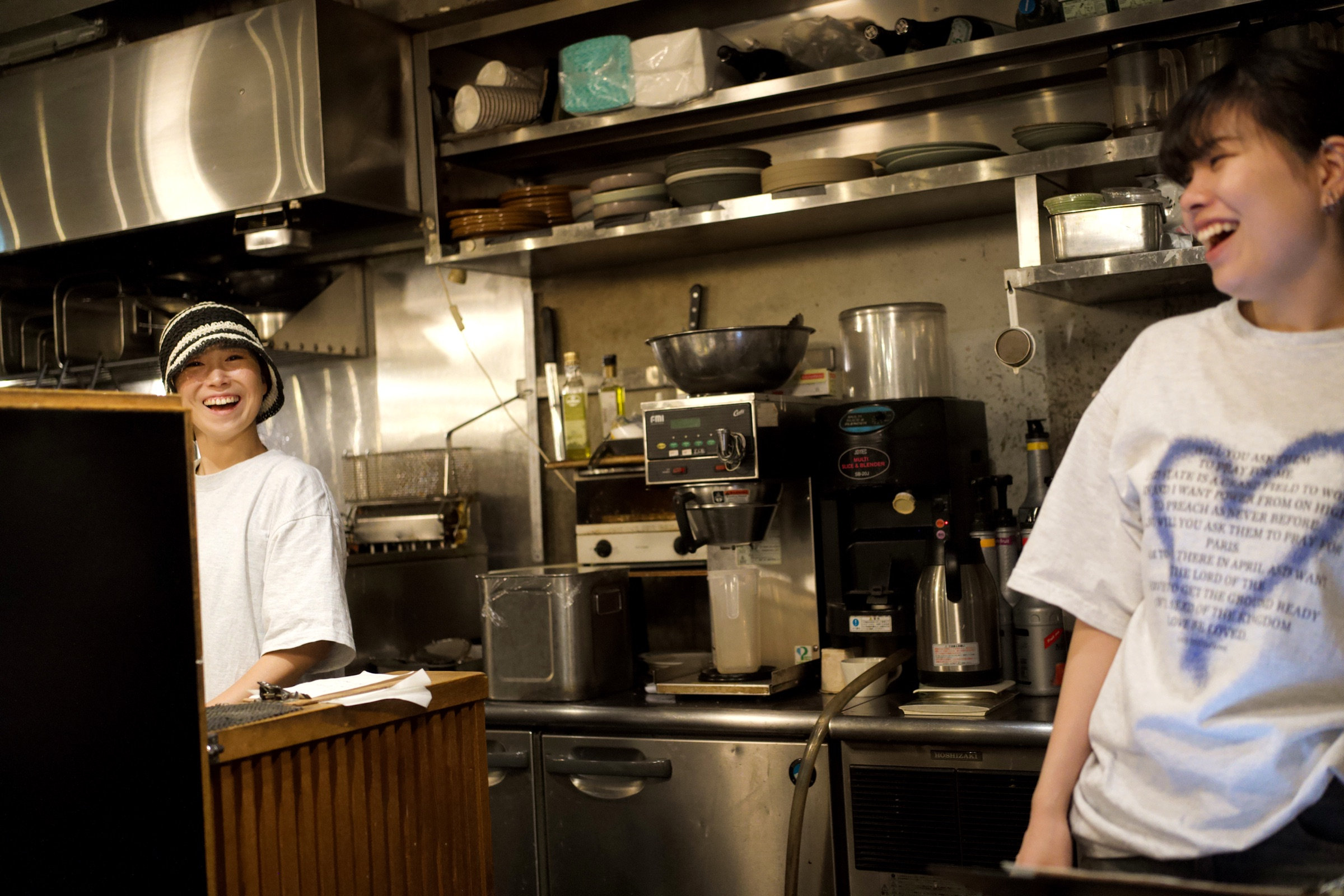 Two women laughing hard in a small Osaka kitchen, one behind the counter in a striped beanie and chef whites