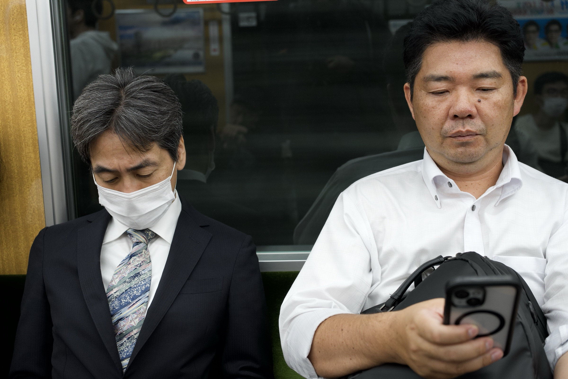 Two salarymen doze side by side on an Osaka train, one in a suit and mask with chin dropping, the other clutching a phone and bag, both surrendered to exhaustion