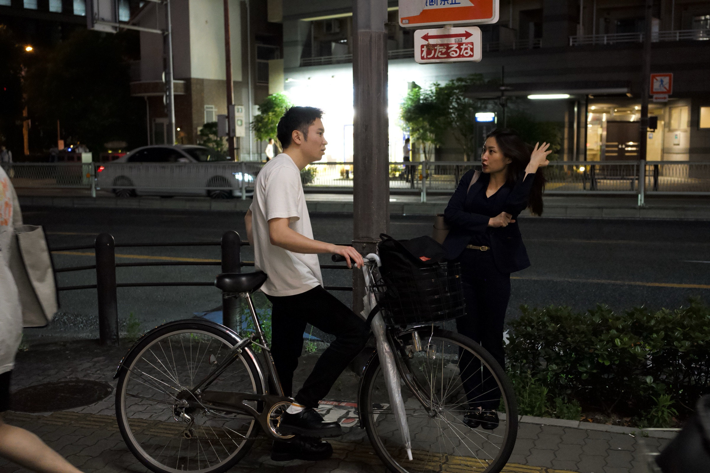 Man on a bicycle and a woman with one hand raised face each other on an empty Osaka street corner at night, a red do not cross sign above them