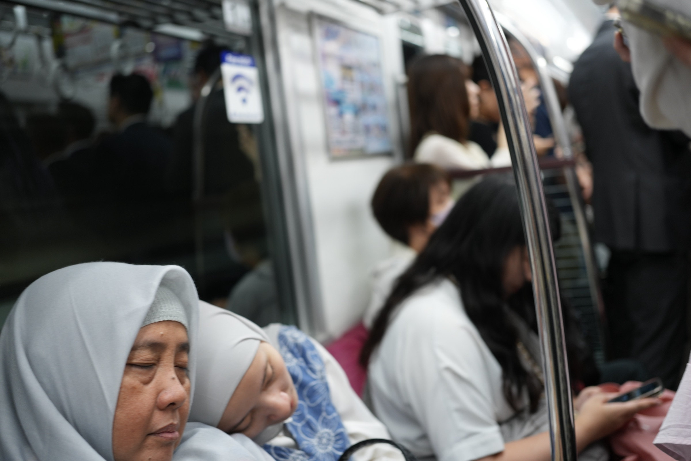 Older woman in a light blue hijab rests on an Osaka subway, a sleeping child nestled against her, other passengers visible through the chrome pole divider