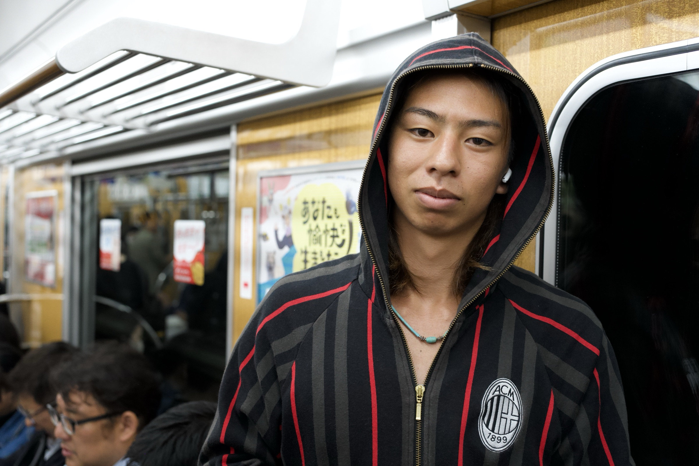 Young man wearing a striped black-and-red AC Milan hoodie with the hood up and wireless earbuds stands in a Japanese train car, looking directly into the camera with a calm expression, advertisements and a seated passenger visible behind him