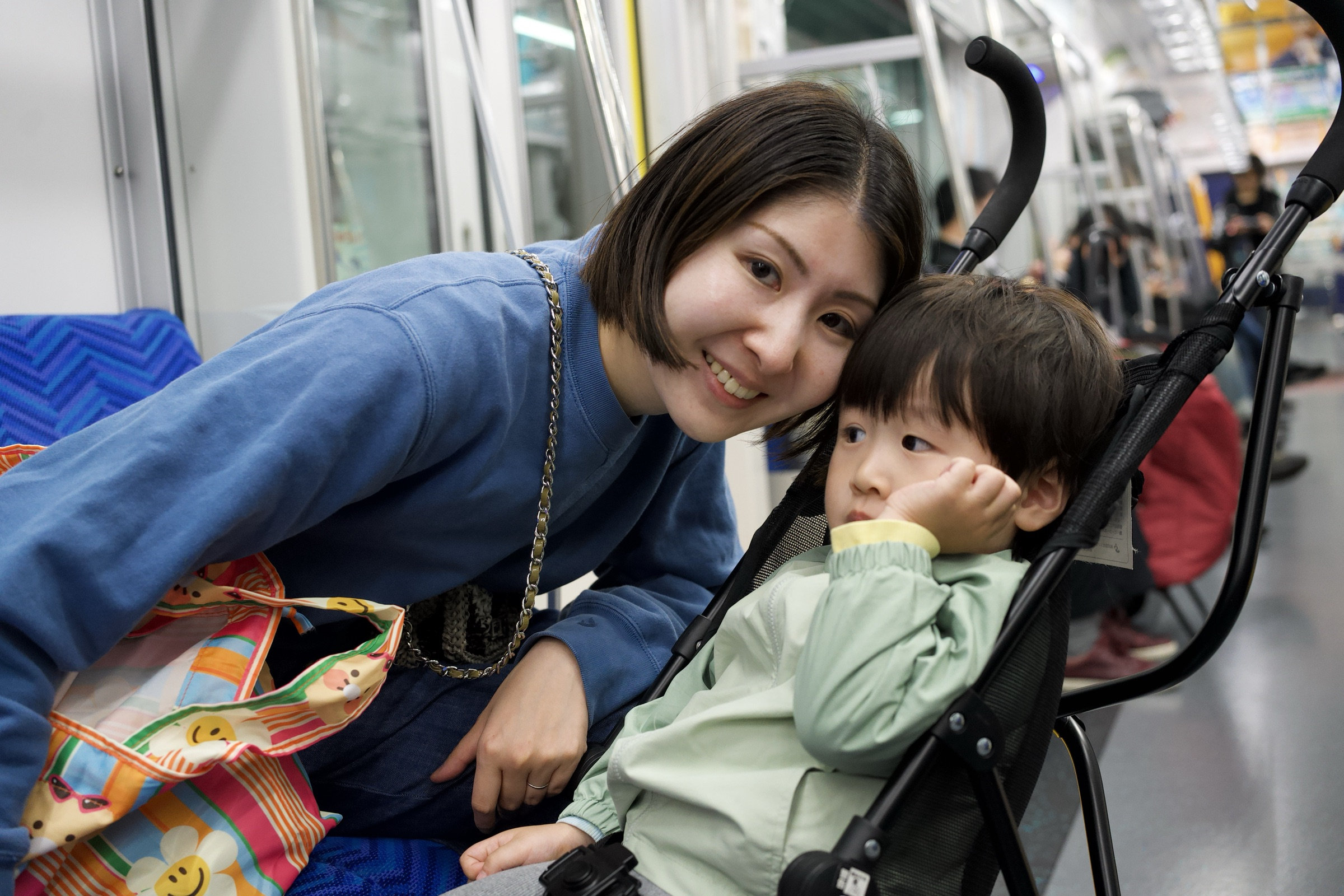 Mother leans cheek-to-cheek with her toddler in a stroller on a Tokyo train, her blue sweatshirt bright against the blue seats, the child's hand on his chin