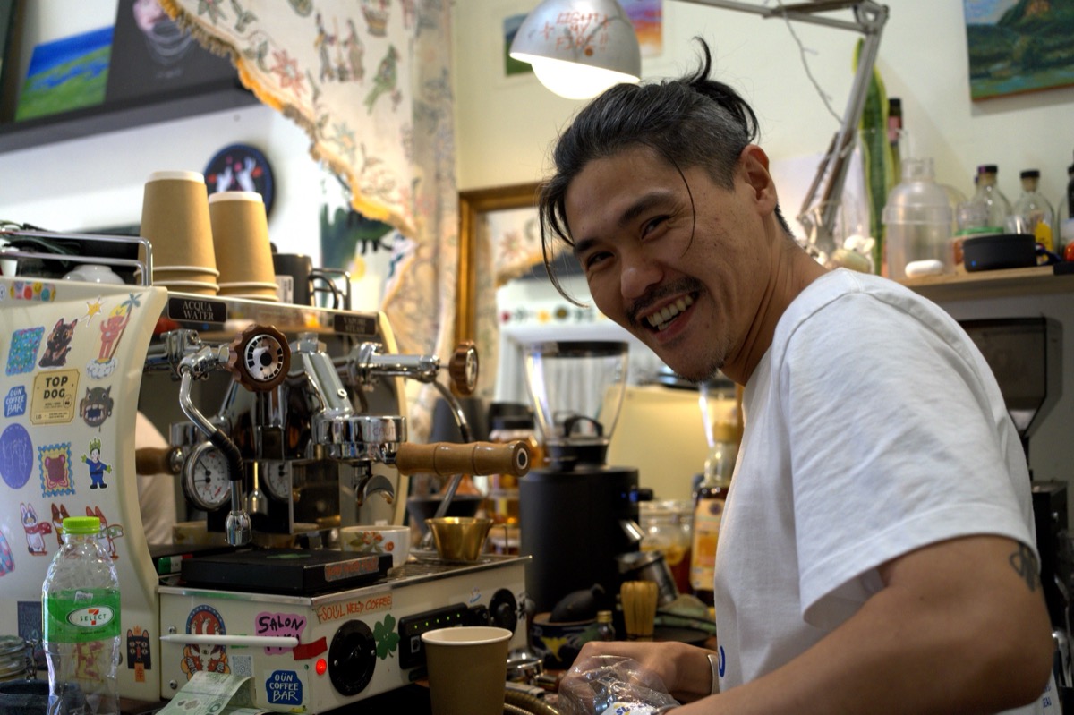 Bangkok barista grinning behind a sticker-covered espresso machine at Soul Need Coffee