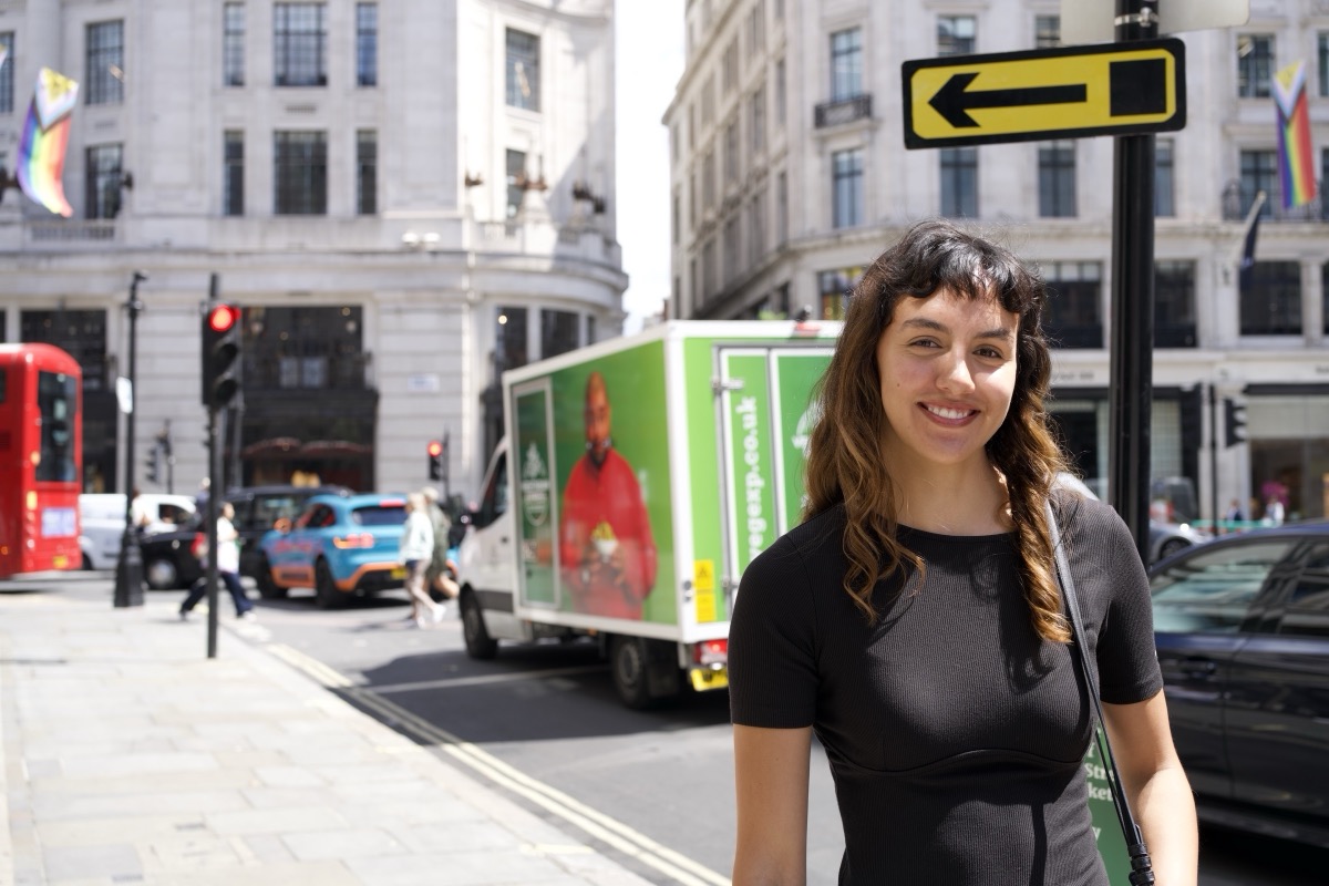 Woman with dark curly hair smiles brightly on a sunny London street, Pride flags and a red double-decker bus behind her