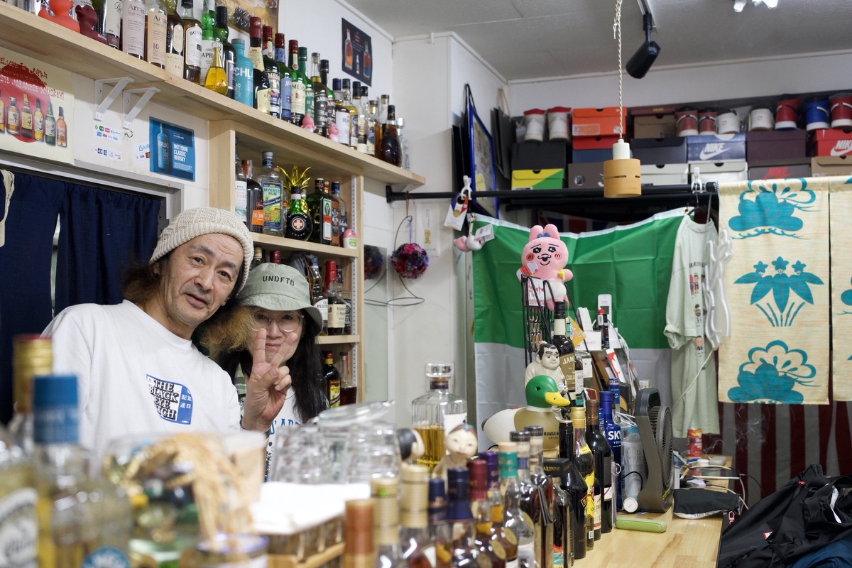 Two Osaka bar owners lean together behind their cluttered counter, bottles and a disco ball behind them